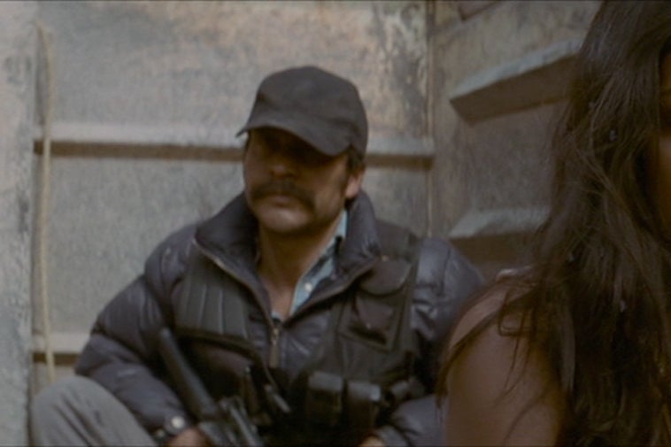 Stephanie Sigman looks down with her face serious and hair unkempt as an armed man dressed in tactical gear watches from behind. 