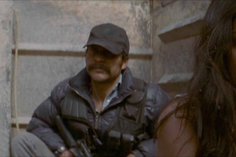 Stephanie Sigman looks down with her face serious and hair unkempt as an armed man dressed in tactical gear watches from behind.