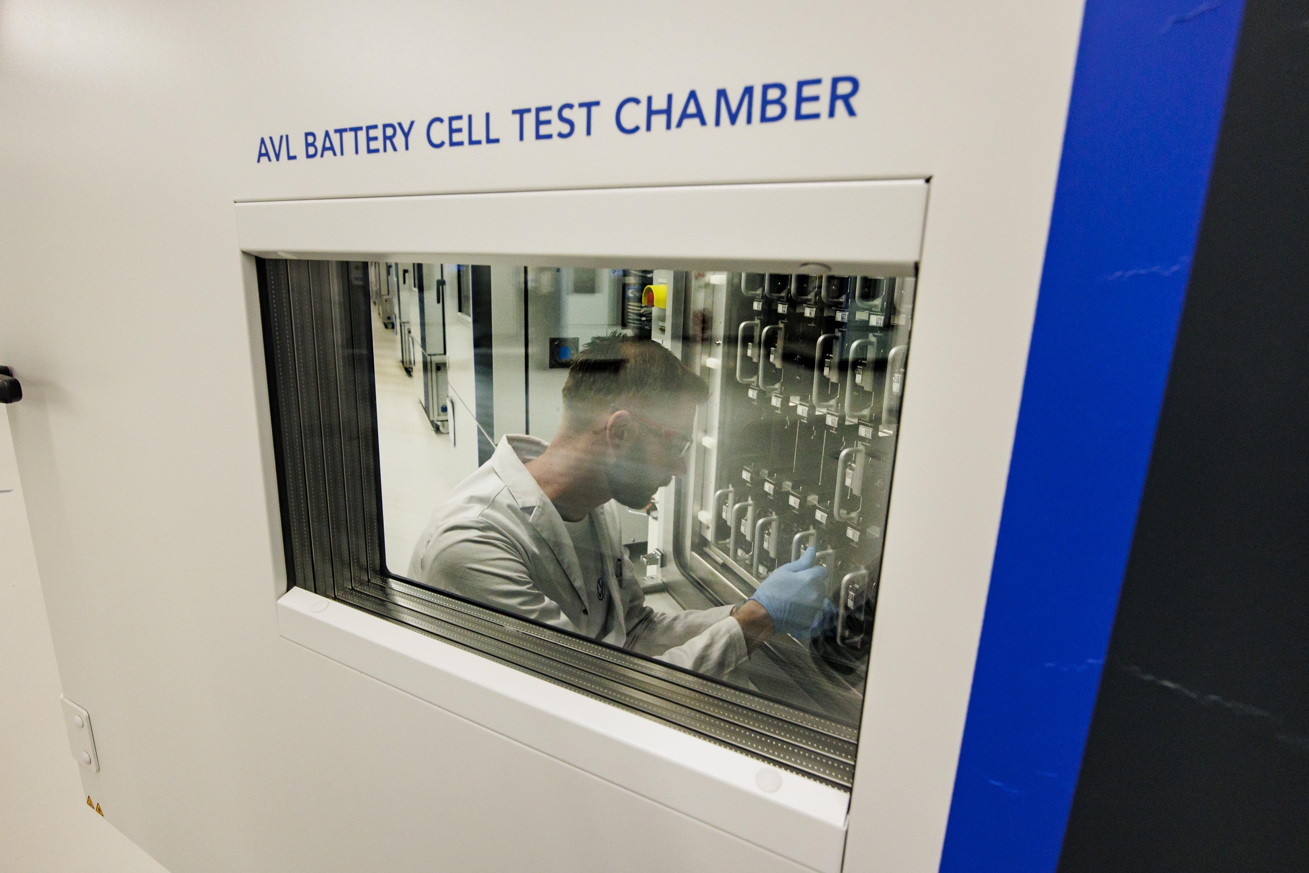 A worker in a lab coat stands behind a door with a window. Above the window, text says "AVL Battery Cell Test Chamber." The worker is working on what appear to be EV car batteries.