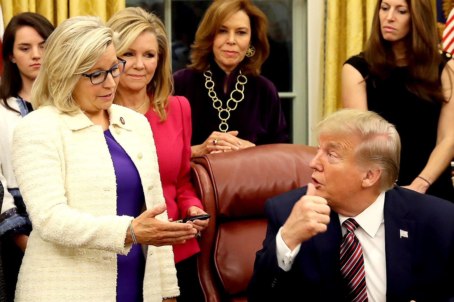 Liz Cheney, standing, speaks to Donald Trump, seated at the Resolute Desk. Several women stand behind them.