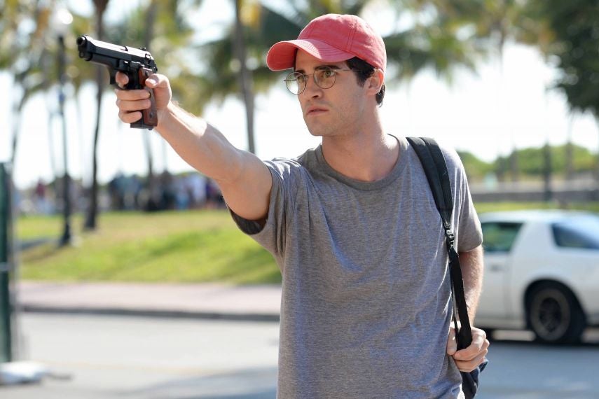 Darren Criss wears a red hat and round glasses and holds up a gun.