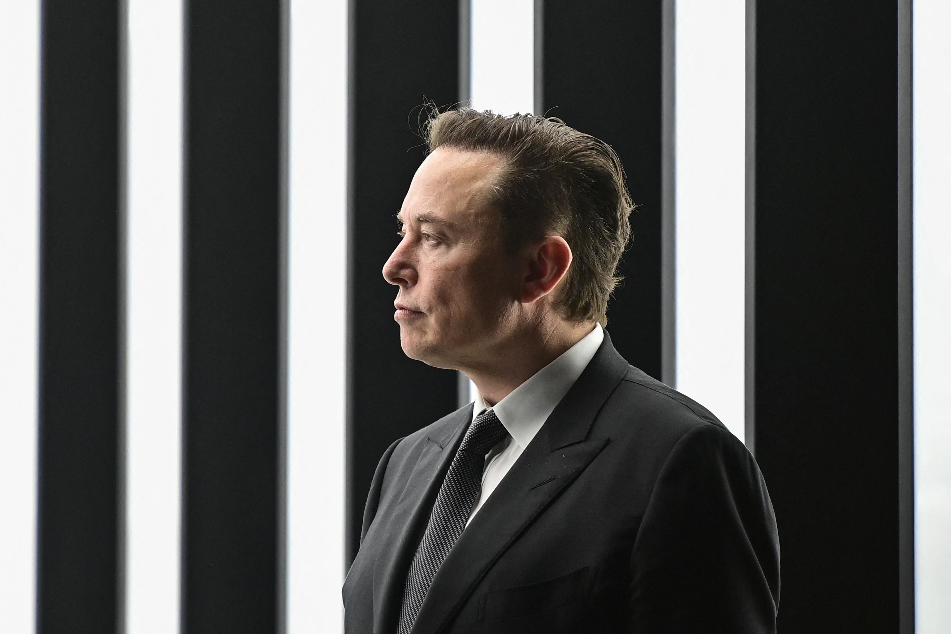 Elon Musk, wearing a suit, stands in front of a background of vertical black and white stripes.