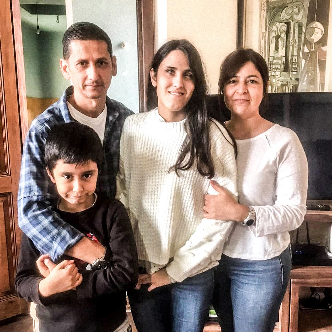 The picture shows a family of four standing in a room with wooden floors and a light green wall. There is a man on the left wearing a blue checkered shirt, and next to him is a young woman in a white sweater. To her right is an older woman, also in a white sweater. In front of them is a young boy in a black shirt and grey pants. The man and the young woman have their arms around the boy. Behind them, there is a wooden door on the left and a framed artwork on the right. They all seem happy and content.