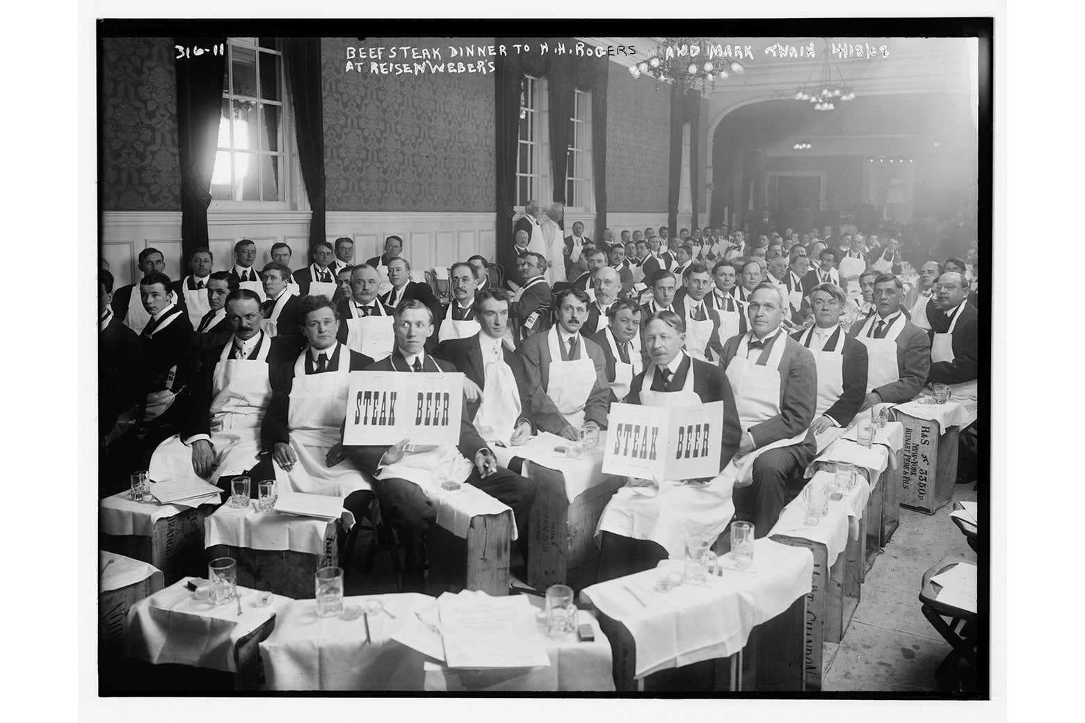 A black-and-white photo of a crowd of men in a restaurant in aprons with signs that say STEAK and BEER.