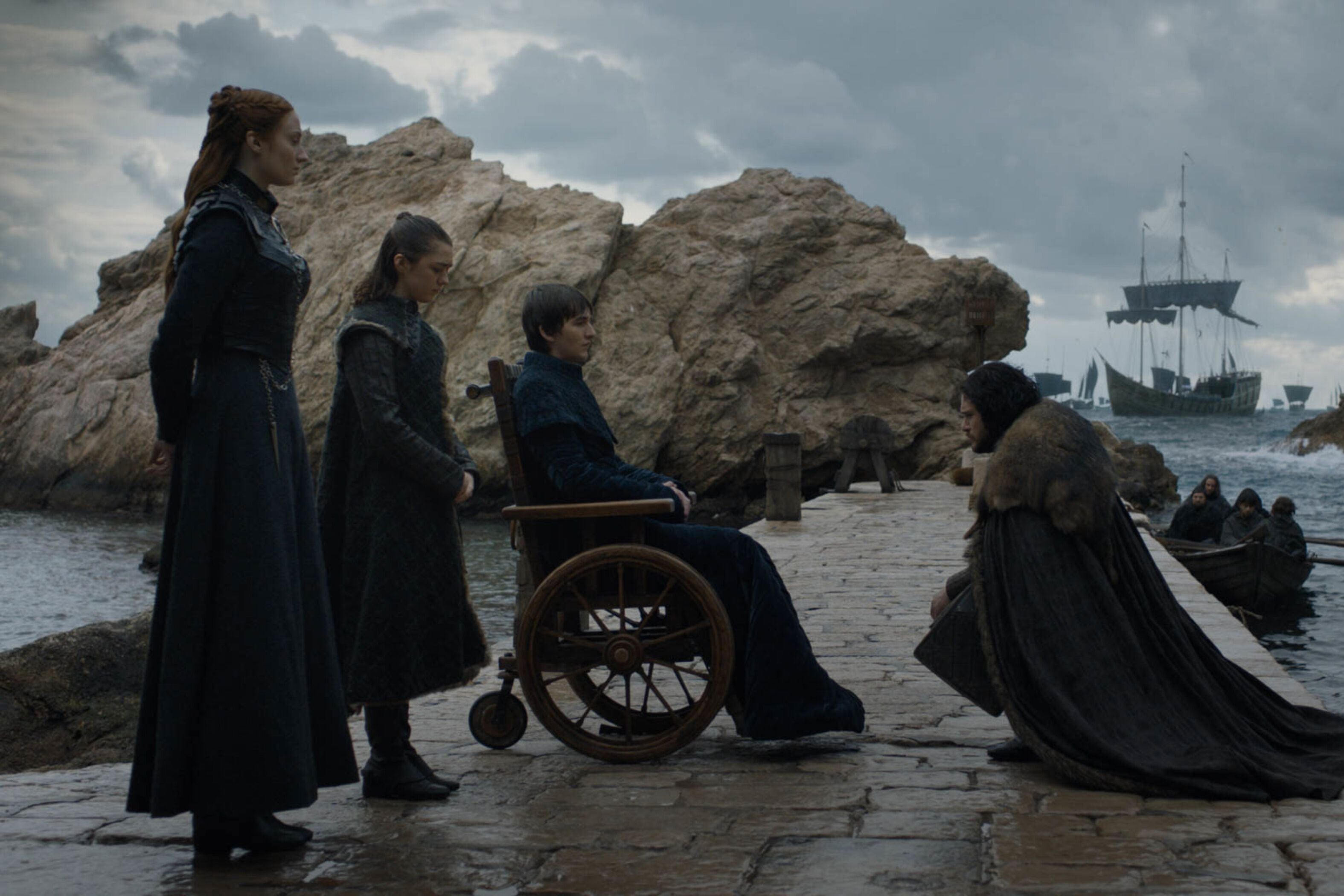 Sansa, Arya, and Bran Stark stand on a jetty with a ship in the background. Jon Snow kneels before them.