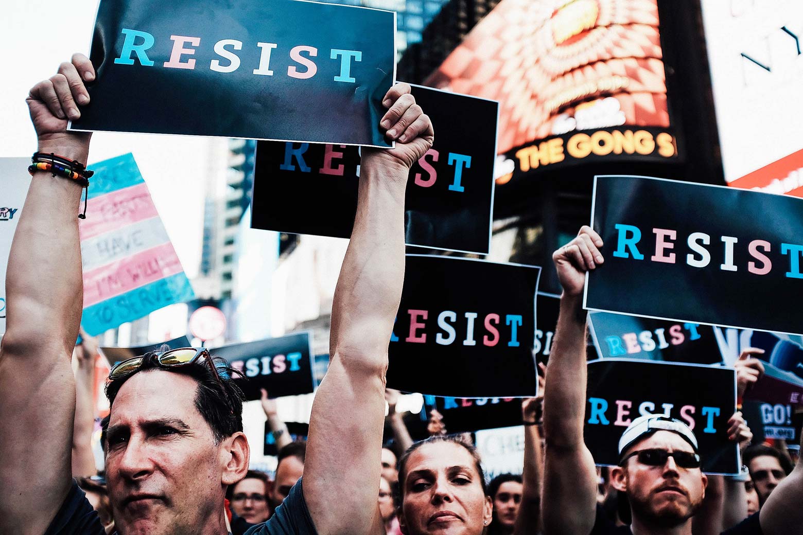 Protesters hold up signs that say, "RESIST" with letters in the blue, white, and pink colors of the transgender symbol.