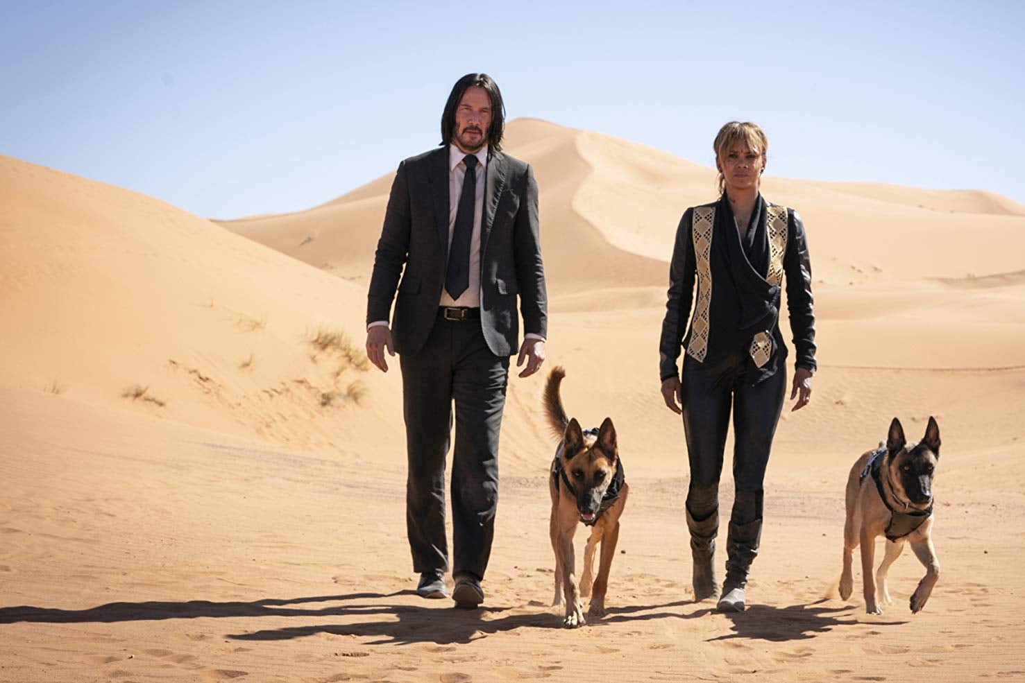 Keanu Reeves, Halle Berry, and two dogs striding across a desert.