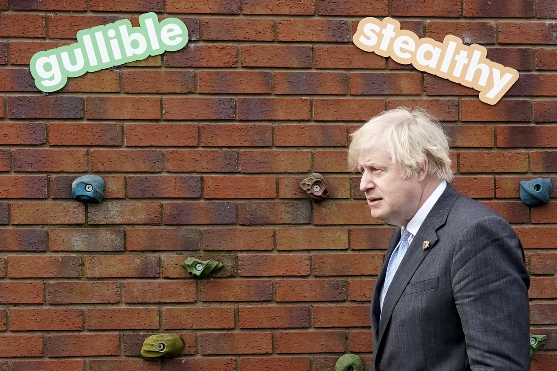 Boris Johnson stands in front of a wall at a school, with two signs reading "Gullible" and "Stealthy" above his head.