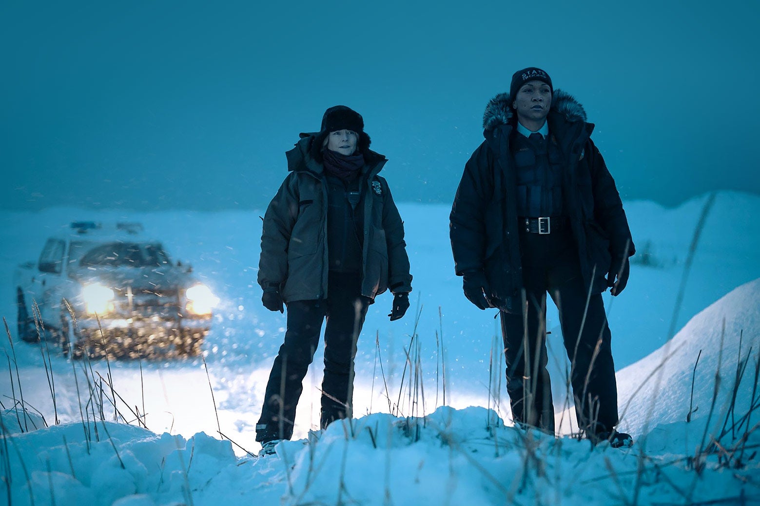 Two female detectives, wearing parkas, stand in the snowy, wintry darkness, with the lights of a police car shining behind them.