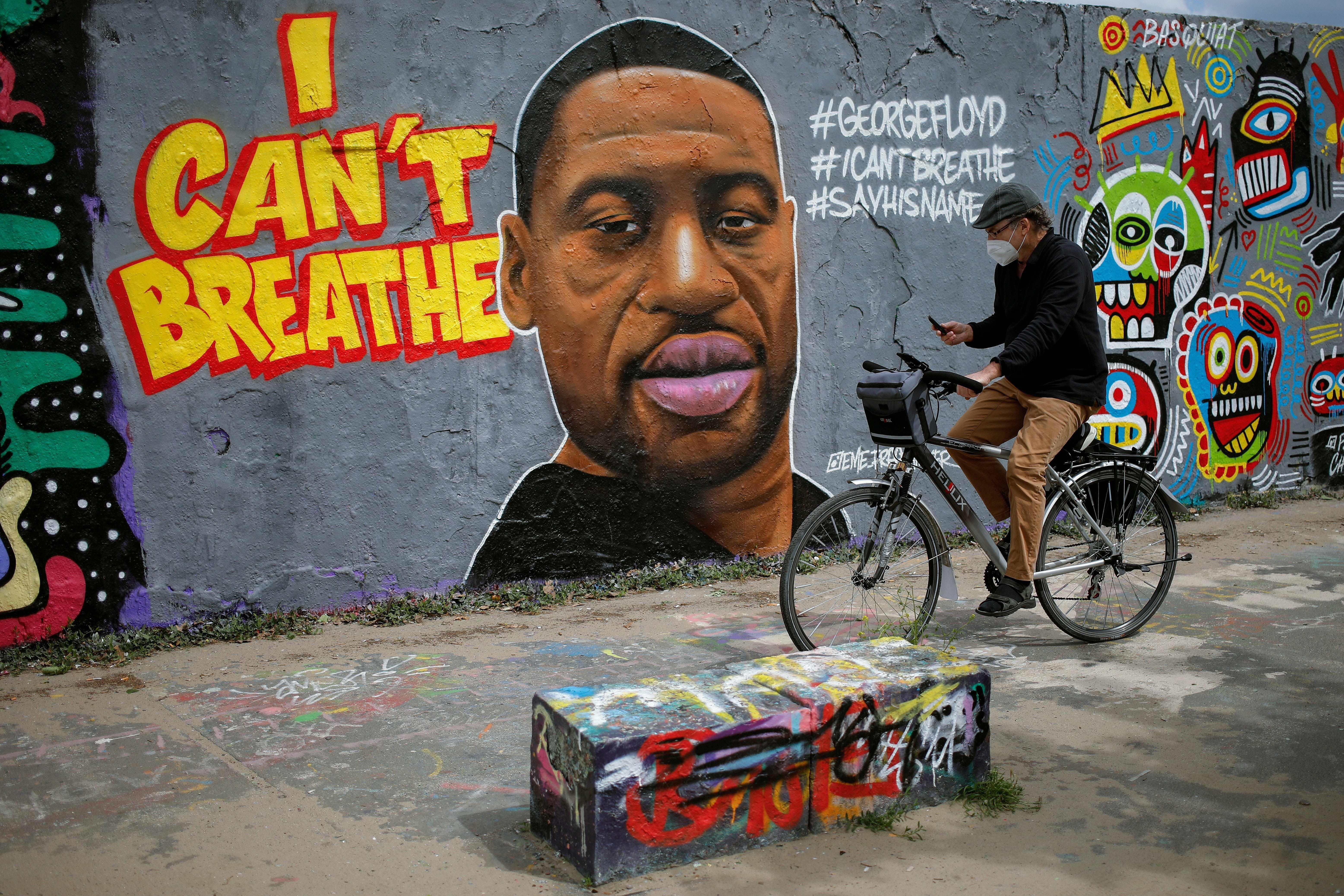 A man wearing a face mask looks at his phone as he cycles passed a graffiti on a wall depicting a portrait of George Floyd, a black man who died in Minneapolis after a white policeman kneeled on his neck for several minutes, is seen on a wall at Mauer Park in Berlin's Prenzlauer Berg district on May 30, 2020. 