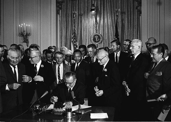 President Lyndon B. Johnson signs the 1964 Civil Rights Act as Martin Luther King, Jr., and others, look on.