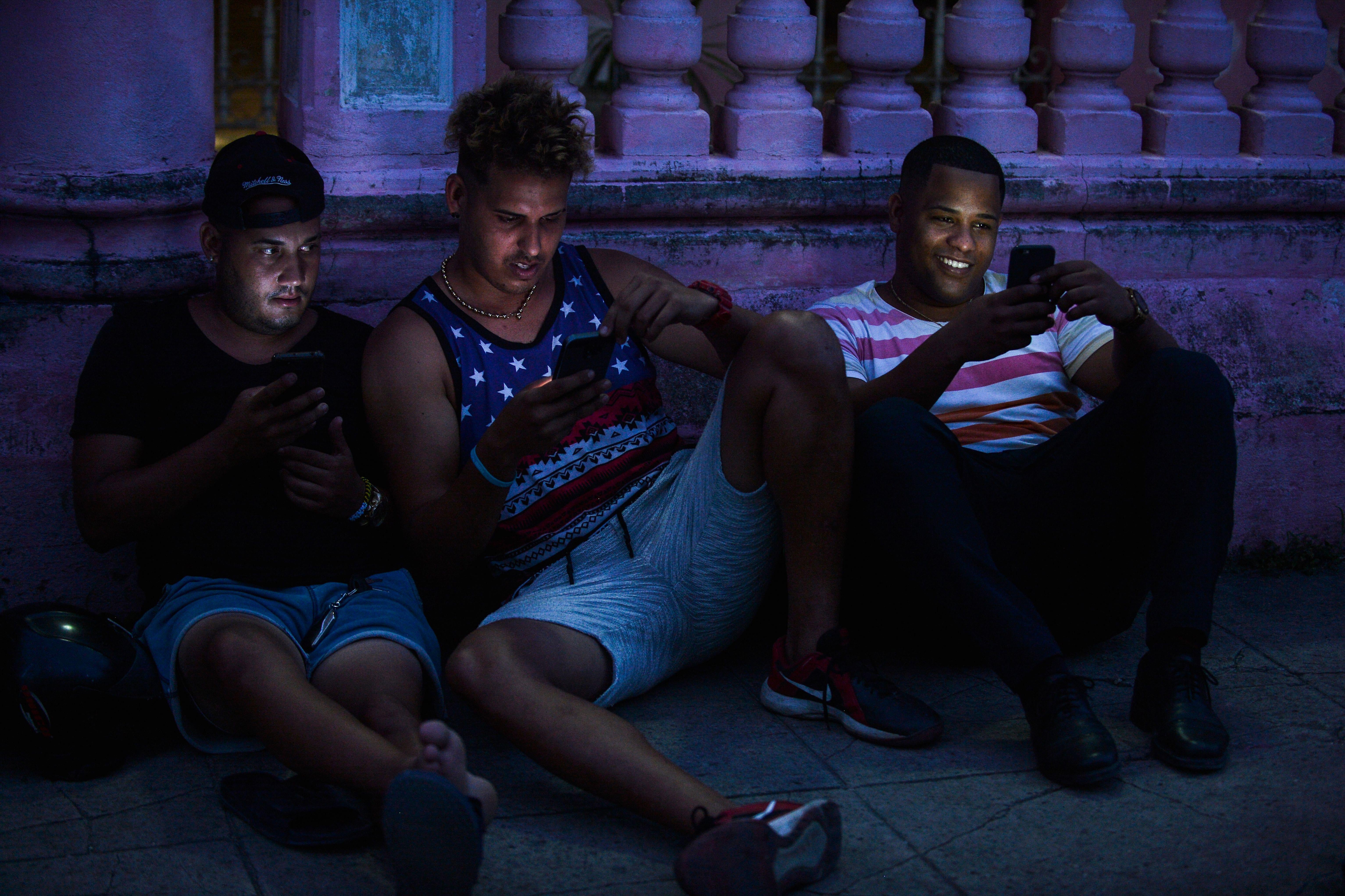Three young men sitting on the sidewalk look at cellphones.