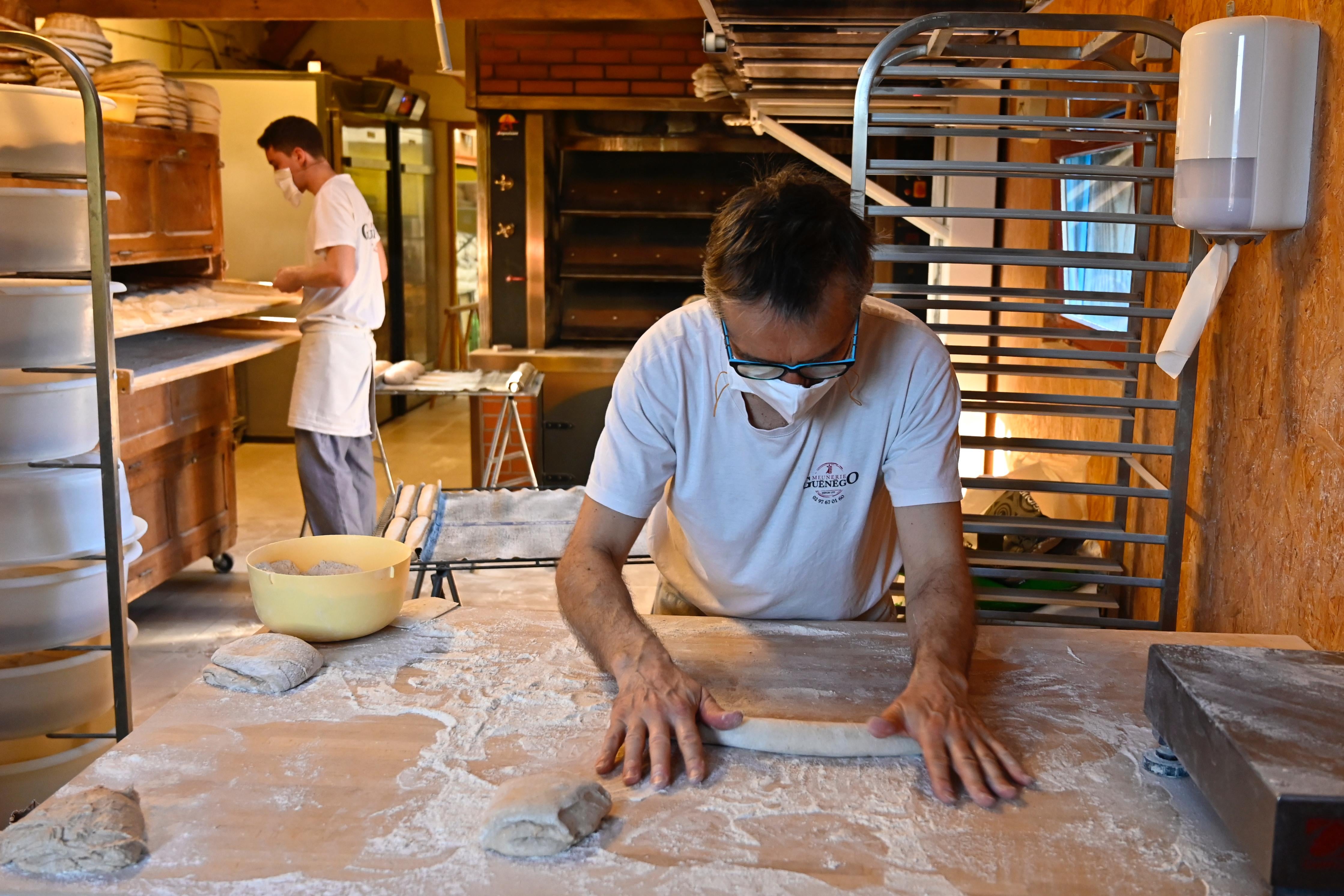 A man in the middle of the frame rolls out dough while wearing a mask. Another man in a mask works behind him.