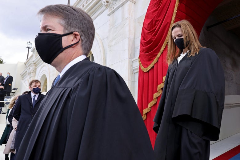 Barrett with her head down wearing a black mask in a black robe following Kavanaugh head up black mask, black robe.