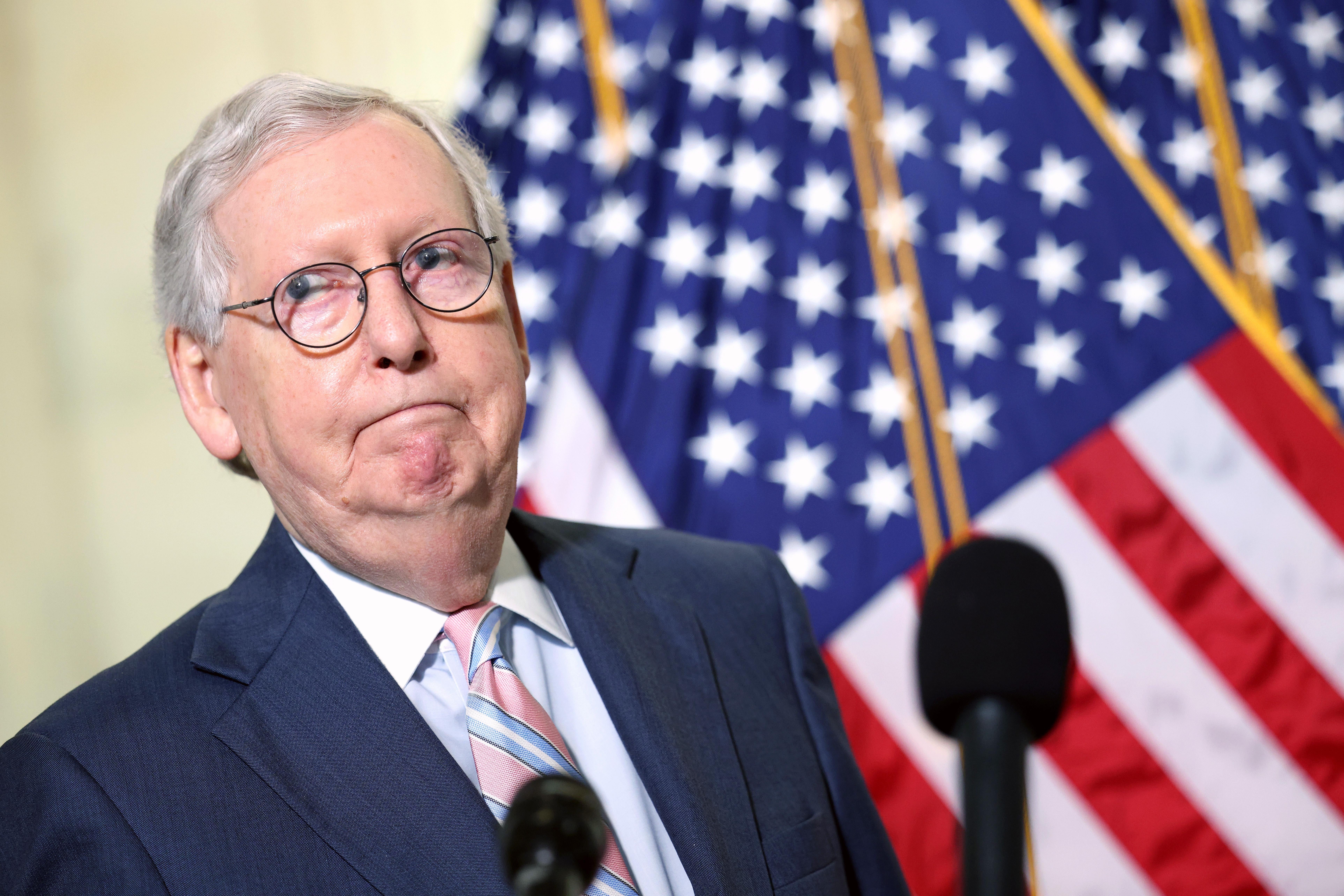 Senate Majority Leader Mitch McConnell frowning in front of a microphone and an American flag.