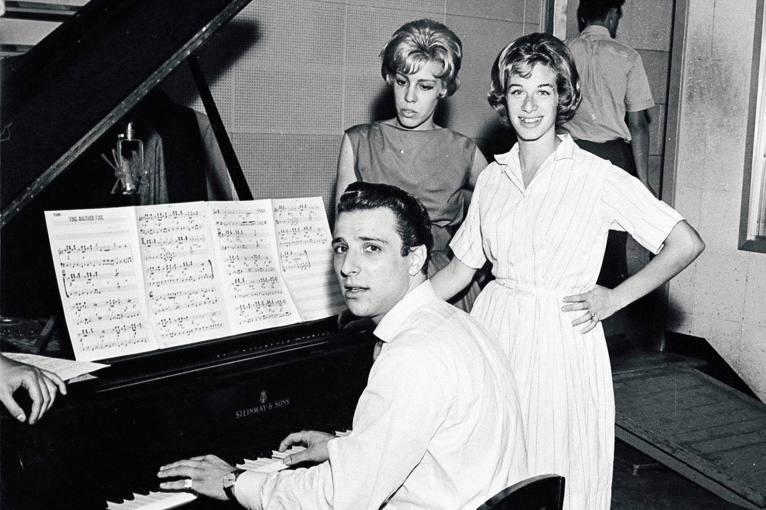 Barry Mann sitting at the piano, with Cynthia Weil and Carole King standing next to him.