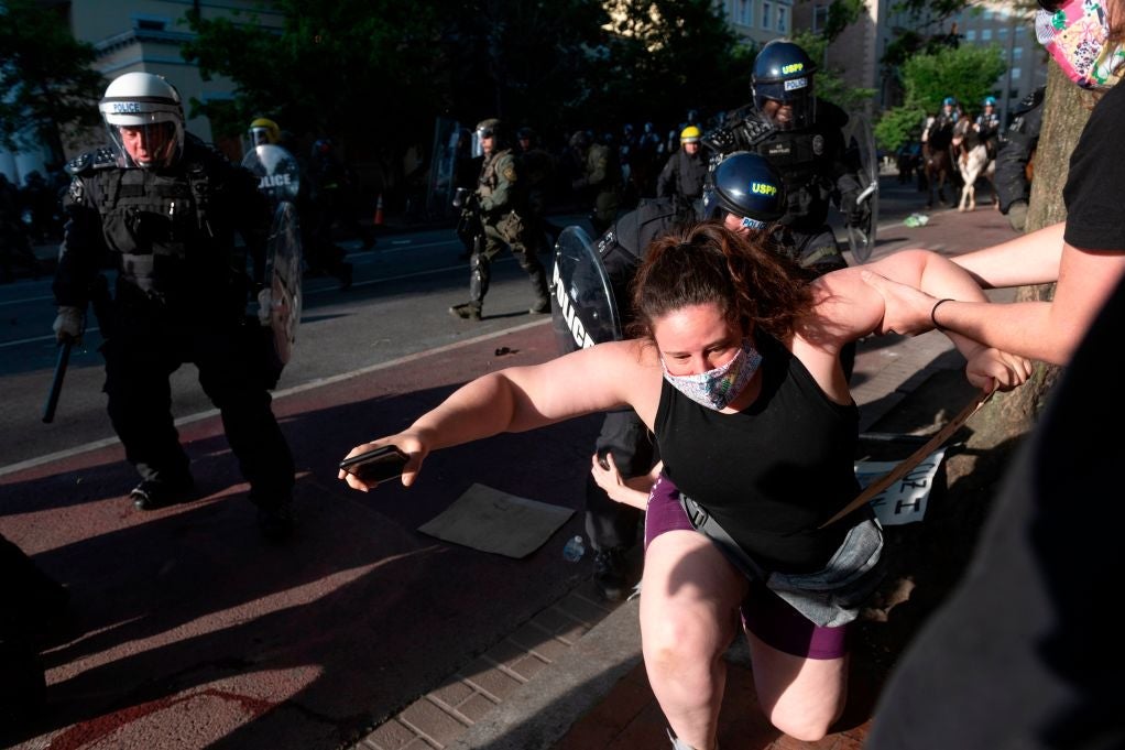 An unassuming woman wearing shorts and a tank top appears to stumble as she is pulled away from onrushing police carrying clubs and shields.