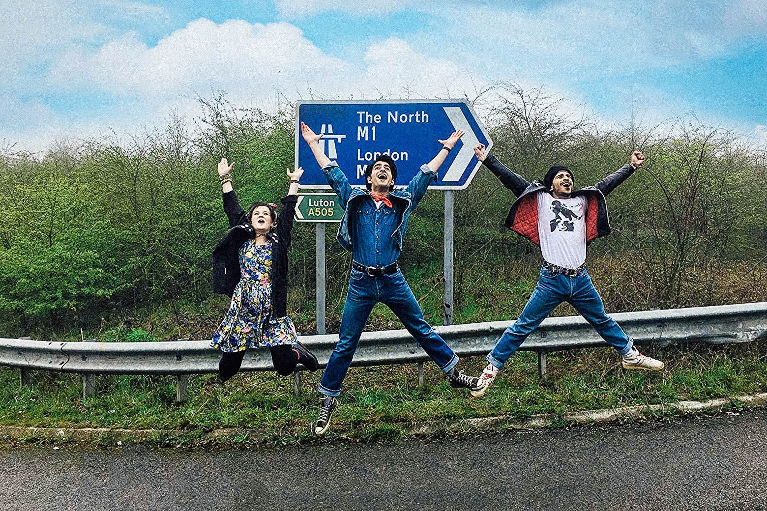 The three characters jump for joy in front of a road sign in a still from the movie.
