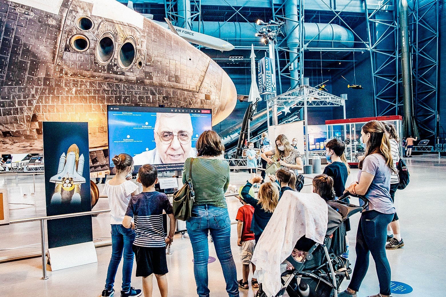 A group of people in a cavernous room with airplanes stand around a screen showing an older man's face.