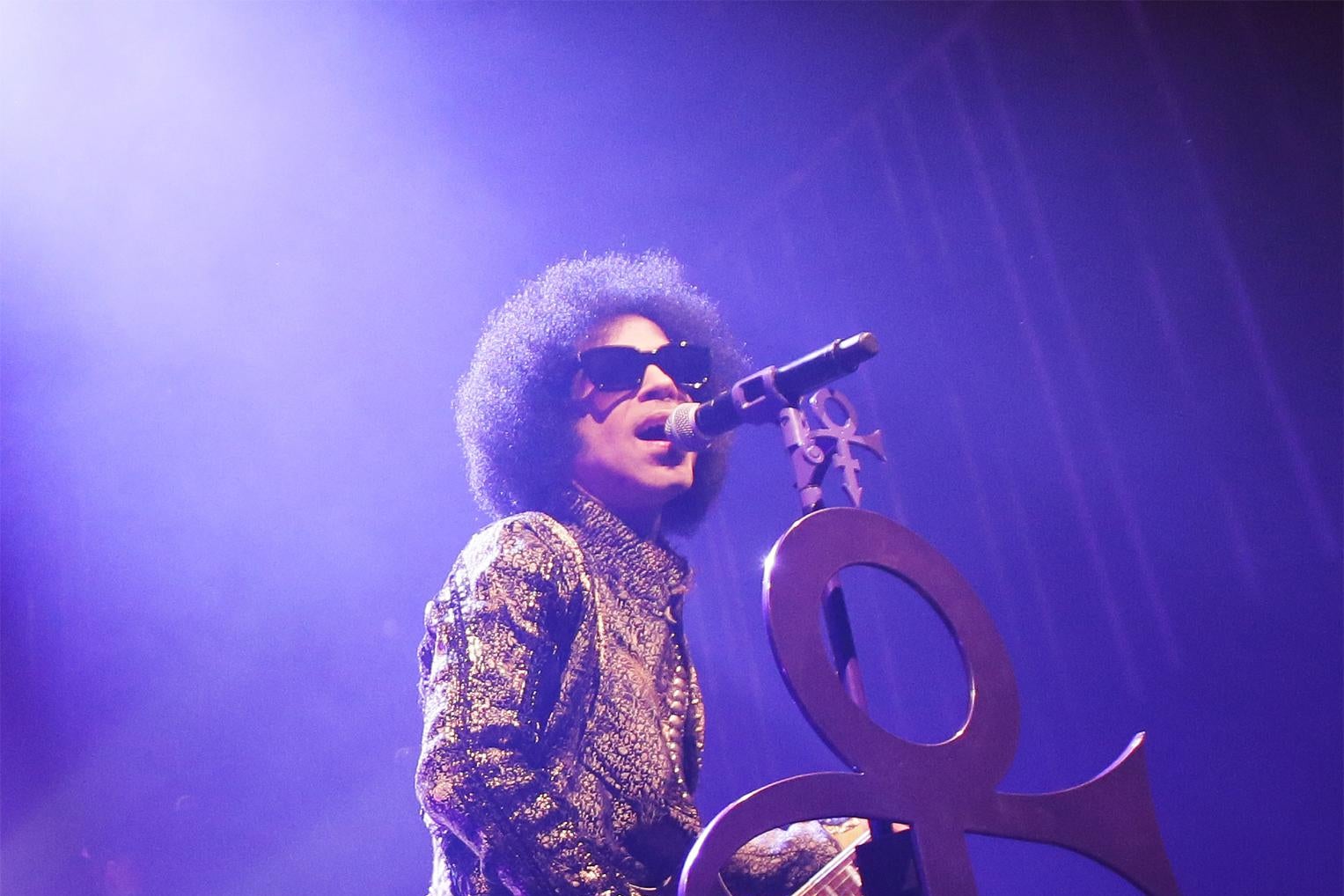 Prince performs in front of a microphone with the Love Symbol, bathed in purple lighting.
