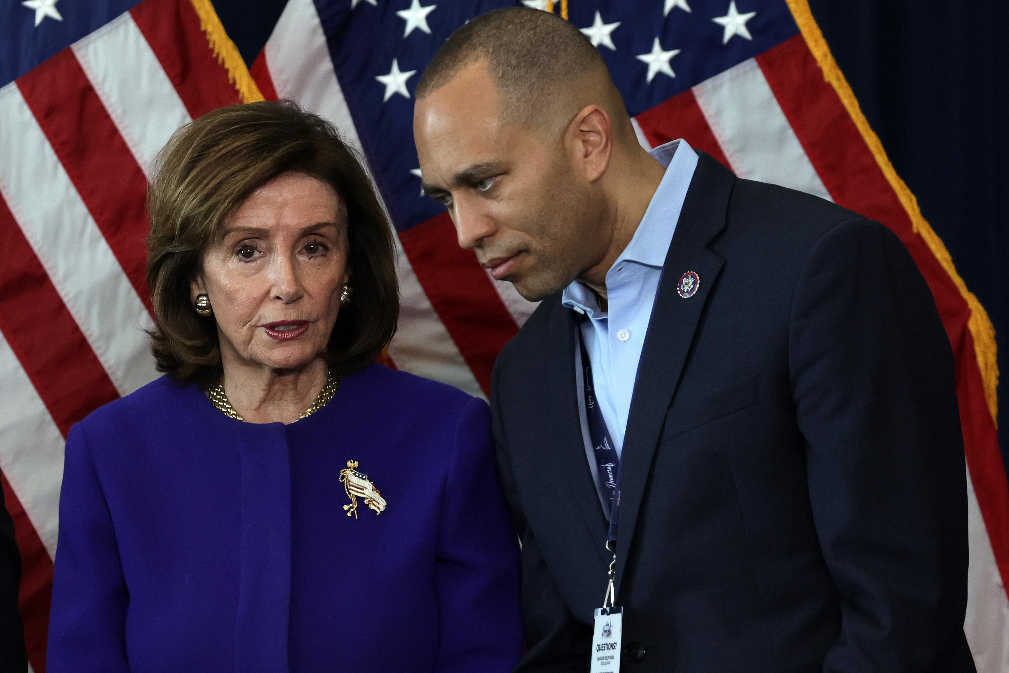 Nancy Pelosi and Hakeem Jeffries lean close to each other, talking, with American flags behind them.
