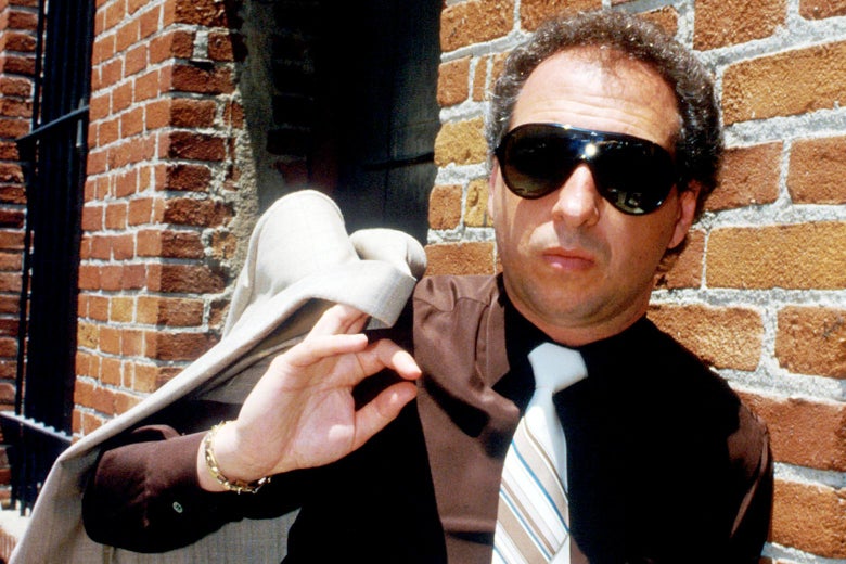 Man wearing large sunglasses and holding a white blazer over his shoulder standing against a brick wall