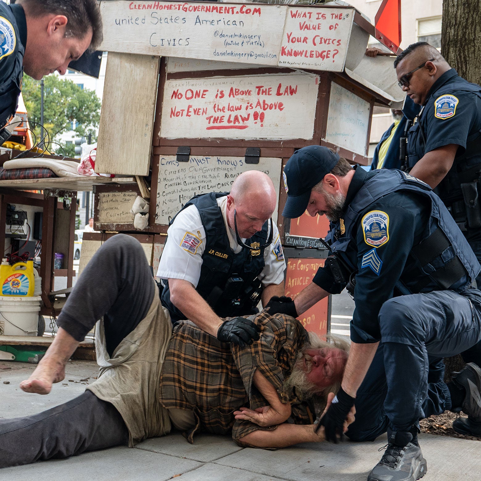 Three officers are holding a barefoot older man down on the sidewalk.