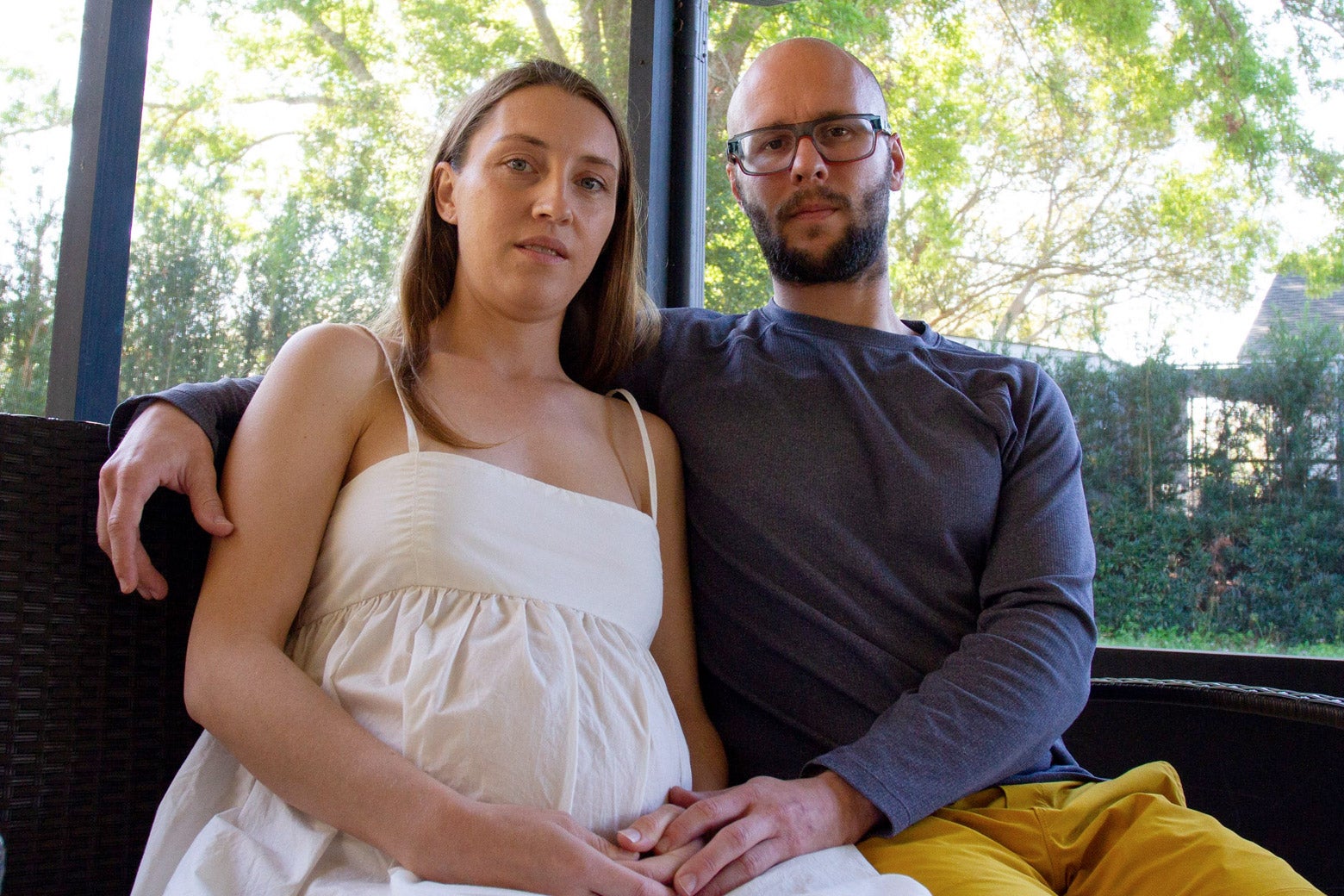 A pregnant woman wearing a white dress sitting next to her husband, wearing a beard, glasses, a black shirt, and yellow pants. Lee looks furious. Deborah looks resigned.