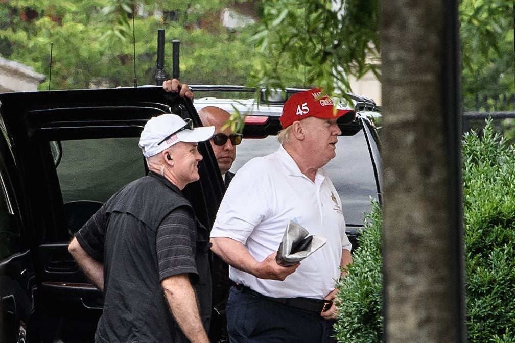 Trump, wearing a white polo shirt and a red hat that says 45, holds a newspaper as he walks from an SUV onto the White House grounds