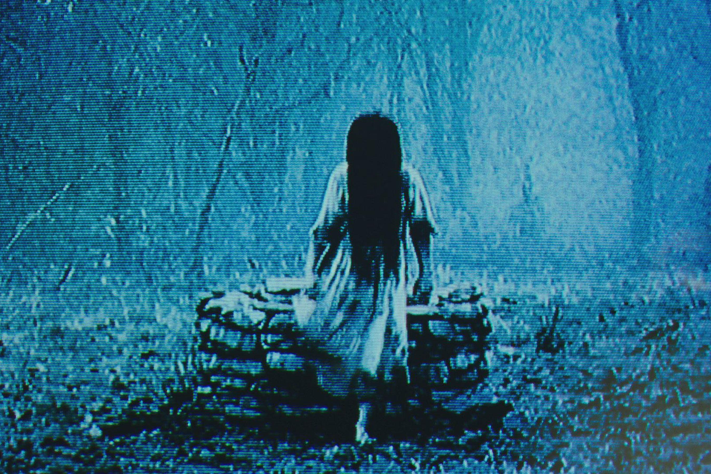 A grainy, blue-tinged image of a girl with long black hair covering her face. She wears a nightgown.