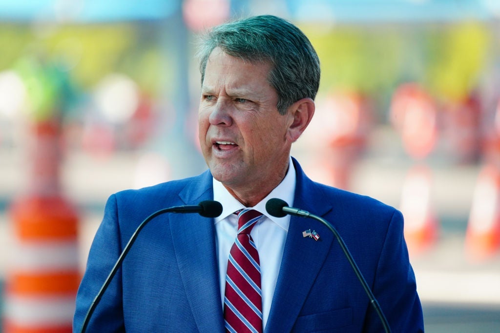 Kemp speaks outdoors at a lectern, wearing a blue suit with a red striped tie.