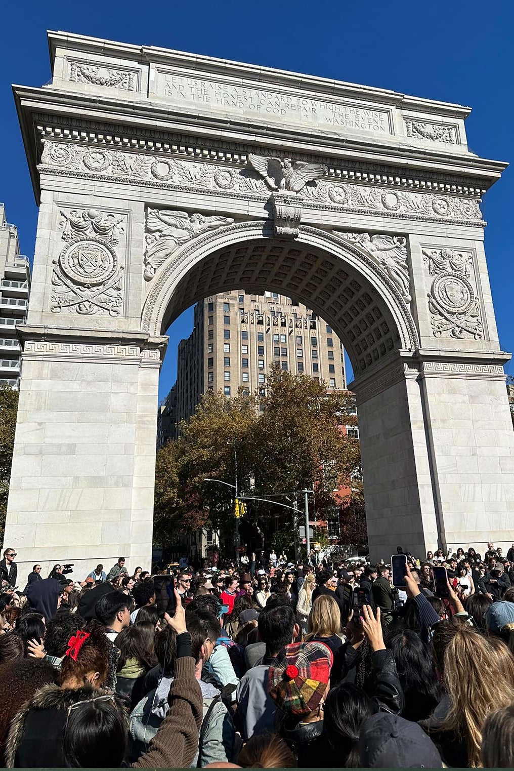 A large crowd of people stands under the famous marble arch in Washington Square Park. They all take out their phones and try to take pictures of the unseen action unfolding in front of them. 