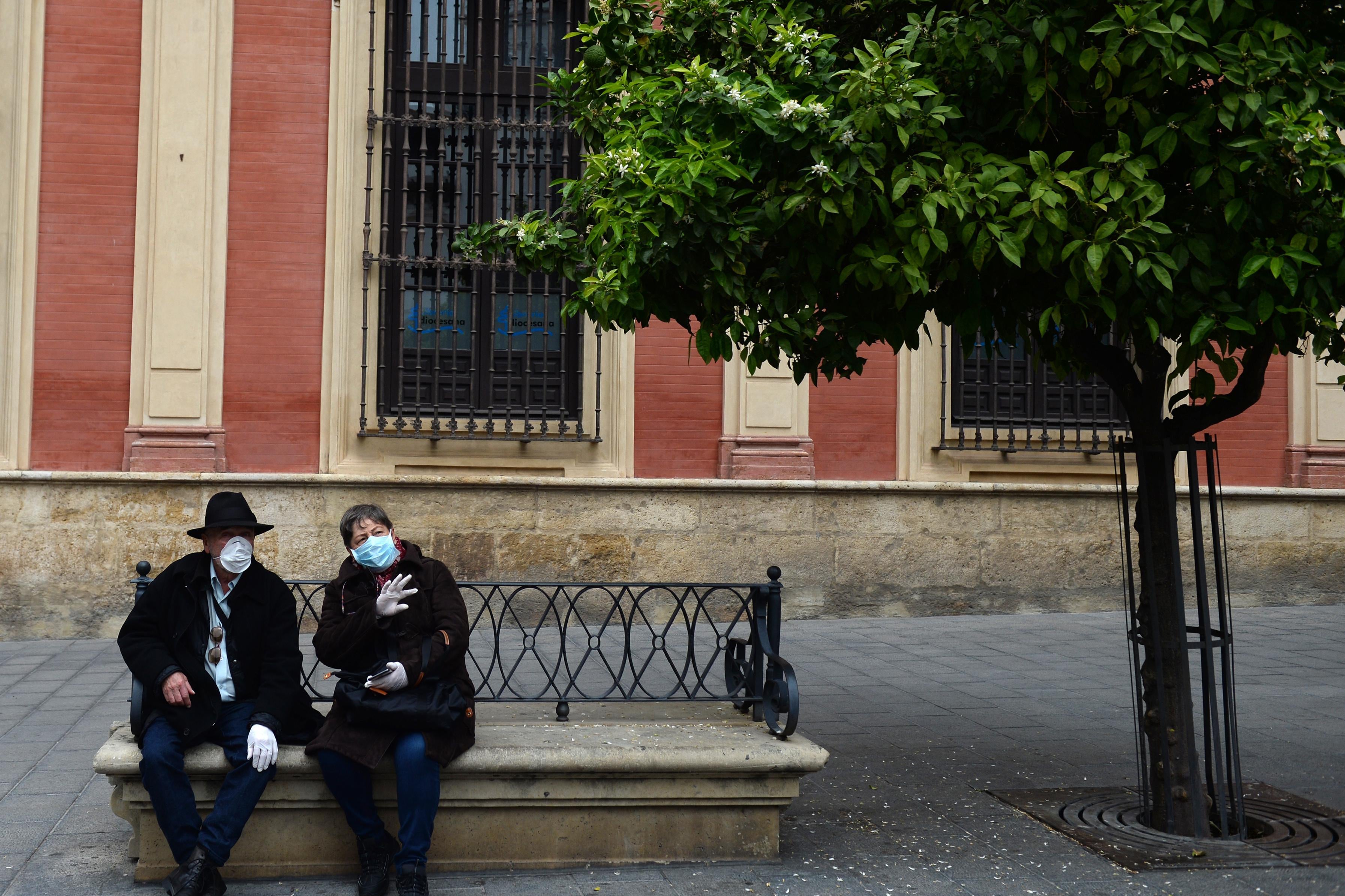 A couple, wearing protective masks and gloves, sit on a bench outside.