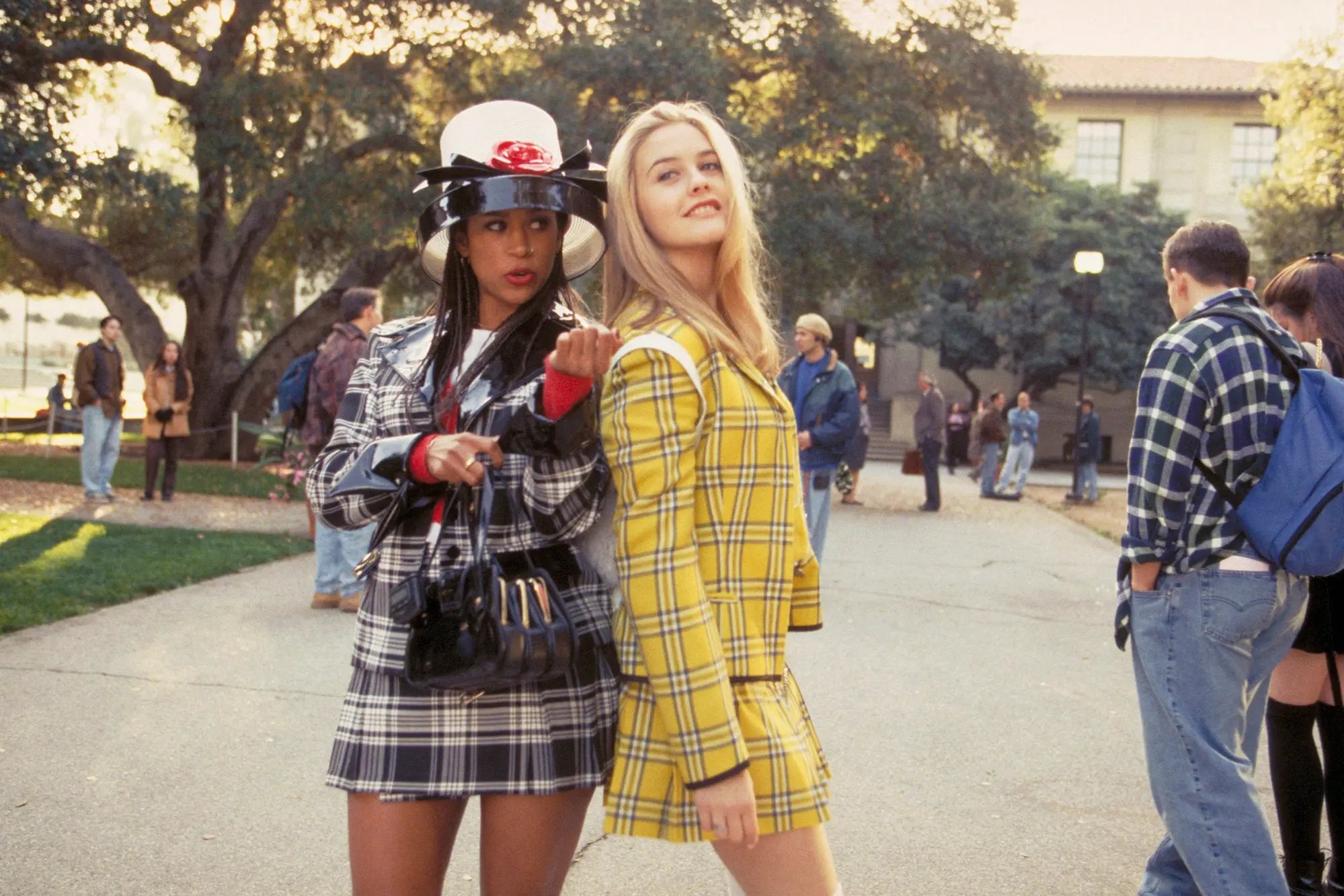 Two girls stand in a high school courtyard. On the left, a Black girl in a preppy matching plaid two-piece skirt and jacket sit and a fancy flashy hat, adorned in stylish accessories. She's talking to the girl on the right, a pretty White blonde in the iconic yellow plaid two-piece skirt set who is leaning on the girl on the left and looking up with a smile on her face.