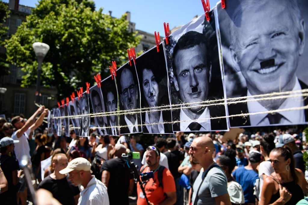 A crowd passes underneath a clothesline pinned with black and white photographs of Emmanuel Macron, Joe Biden, and others, which have been altered to feature "Hitler mustaches"