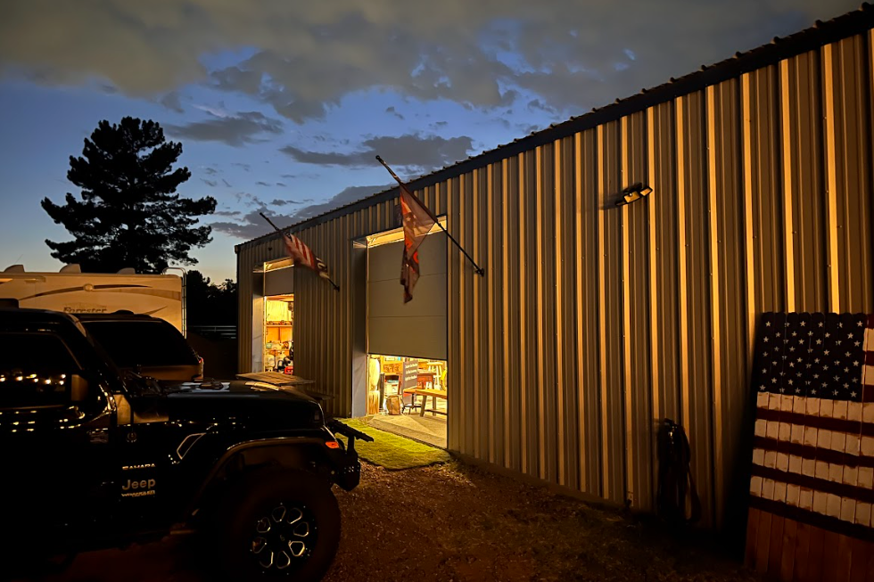 The exterior of the garage, a white metal structure, at dusk. Several American flags hang outside the building, and a number of cars are parked outside.