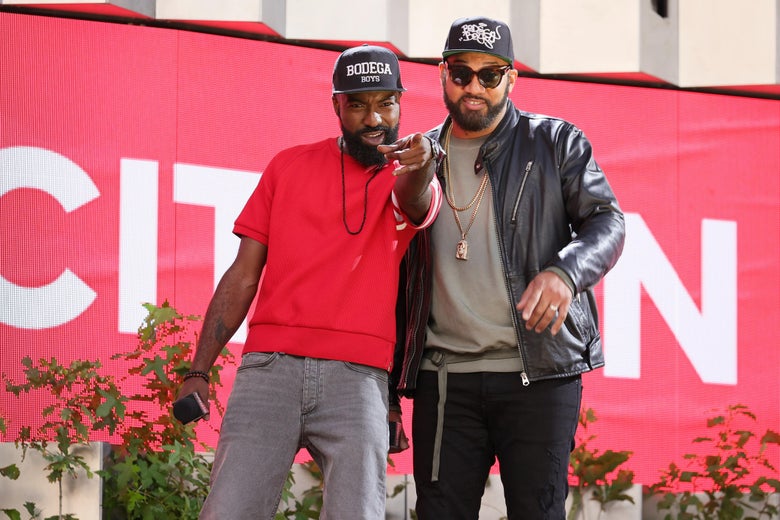 Desus and Mero stand in front of a banner, with Desus pointing at the camera.