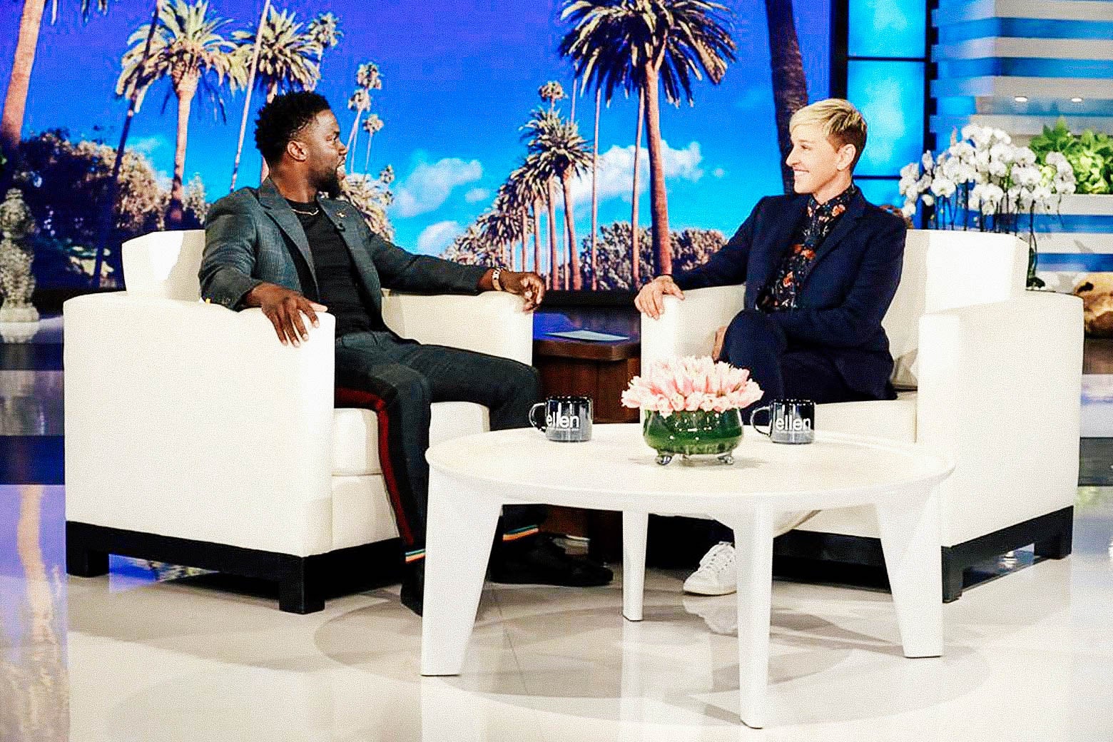 Kevin Hart and Ellen DeGeneres sit in white chairs on the set of her show.
