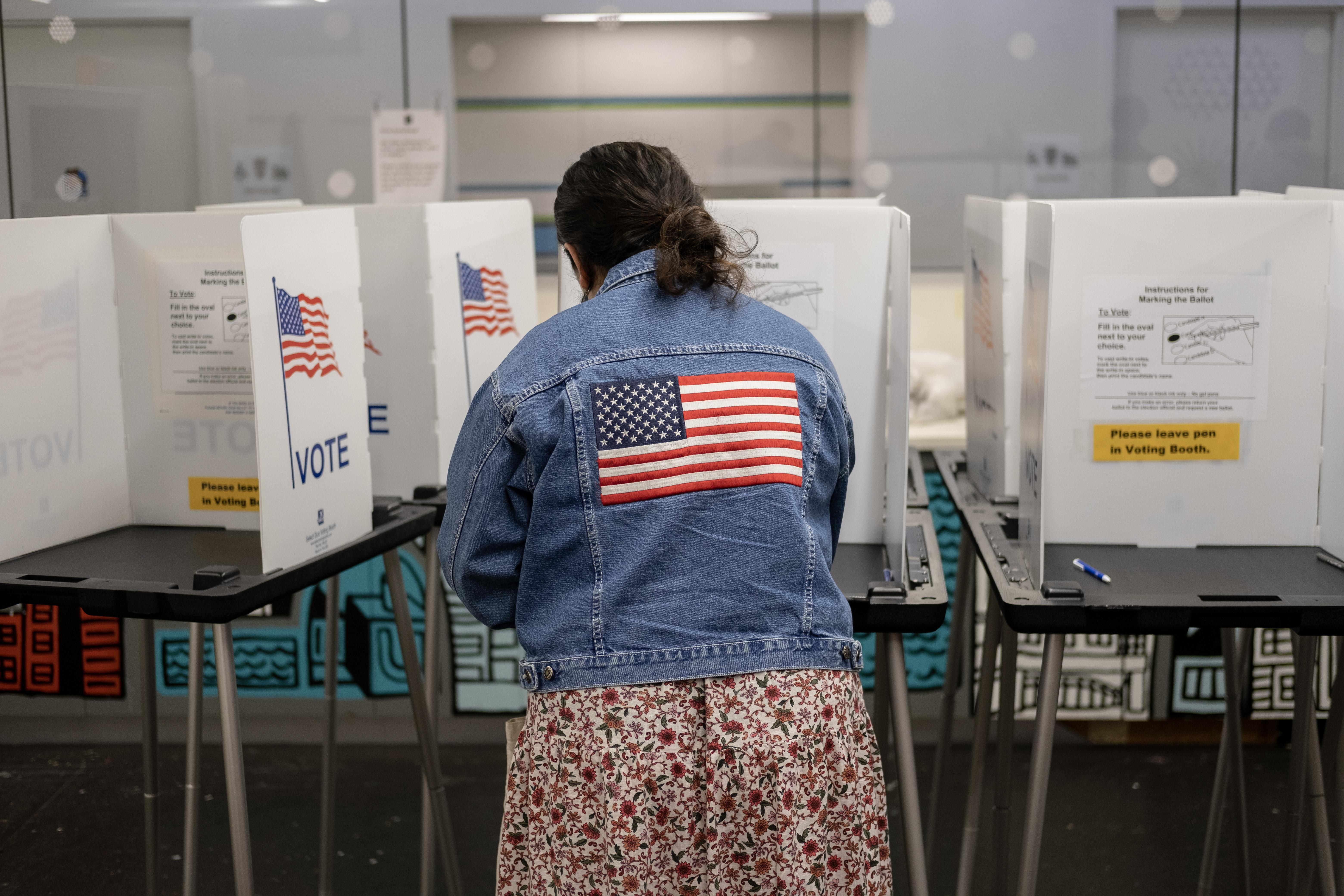 A woman wearing a dress and a jean jacket with an American flag on her back casts her ballot.