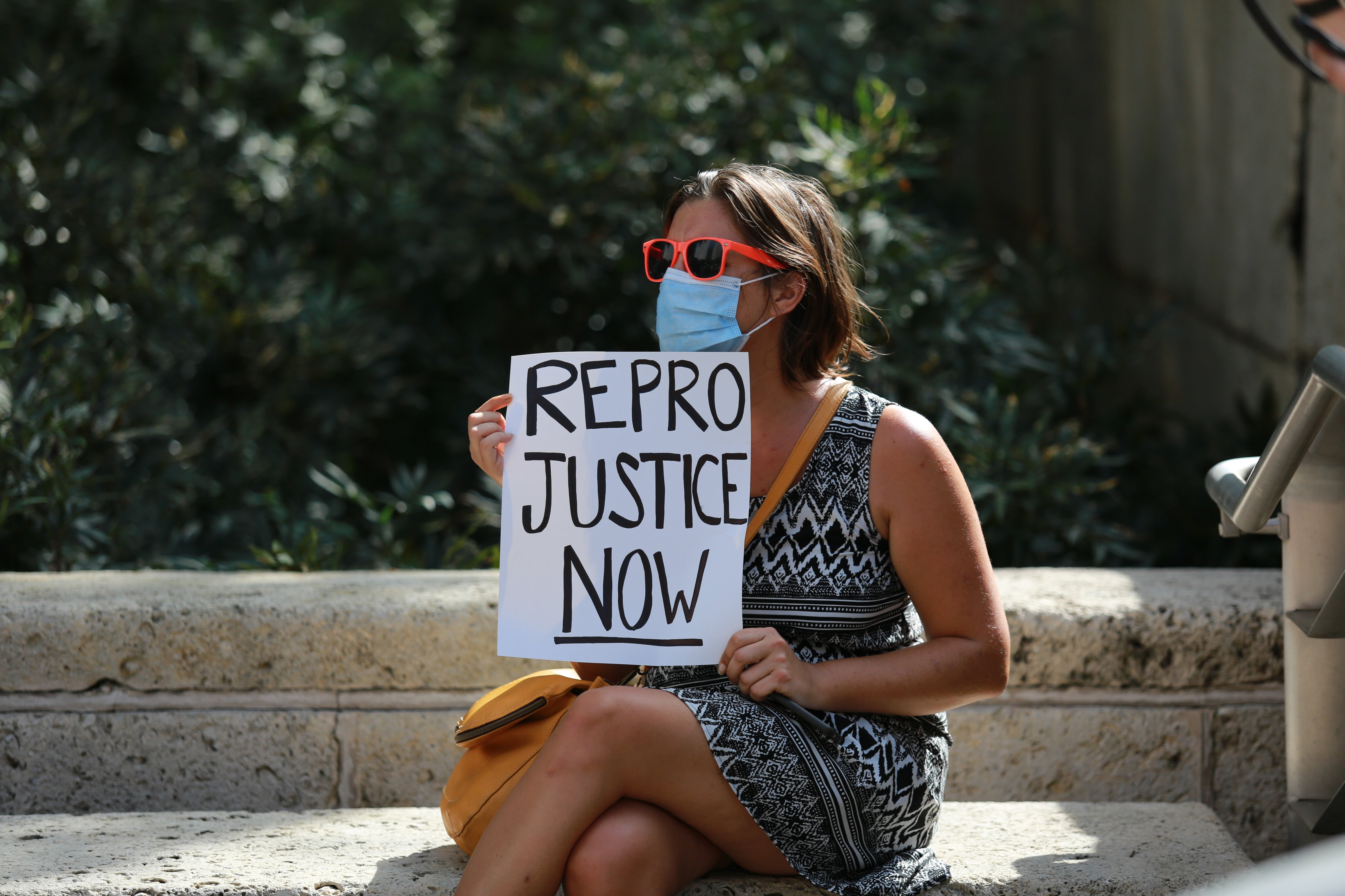 A woman in a dress sits on a bench wearing a mask and sunglasses, holding a sign that says, "REPRO JUSTICE NOW."