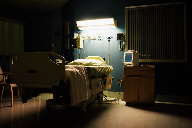 A hospital bed is seen in a patient room under a shining light. To its right side is a cabinet with a monitor on top.