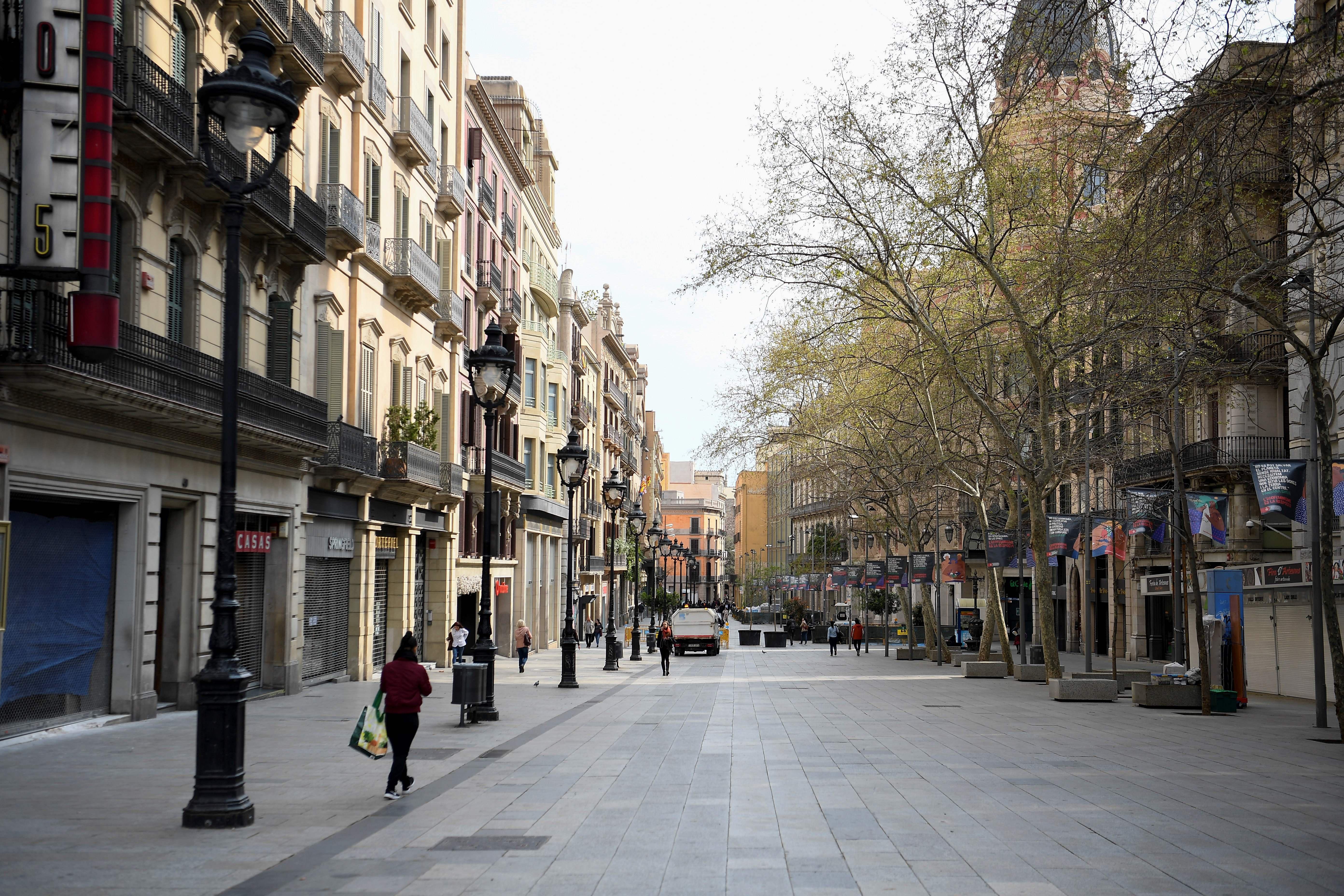 A Barcelona street, lined with shops, nearly empty. A lone woman walks away from the camera with a shopping bag.