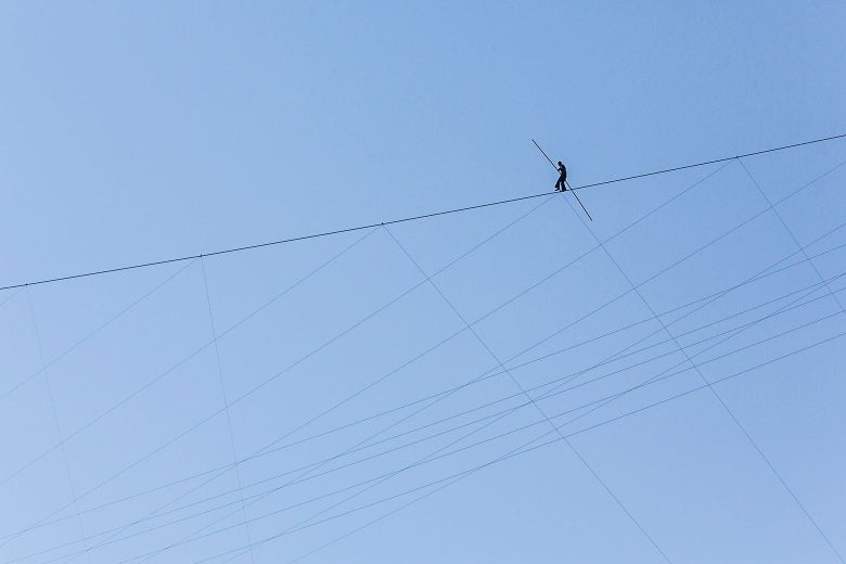 The very small figure of a tightrope walker is seen against a blue sky.