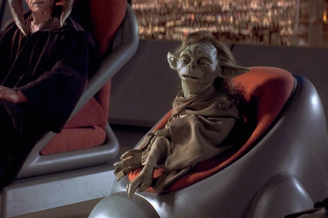 A green, Yoda-like creature with long brown hair sits in a round chair.