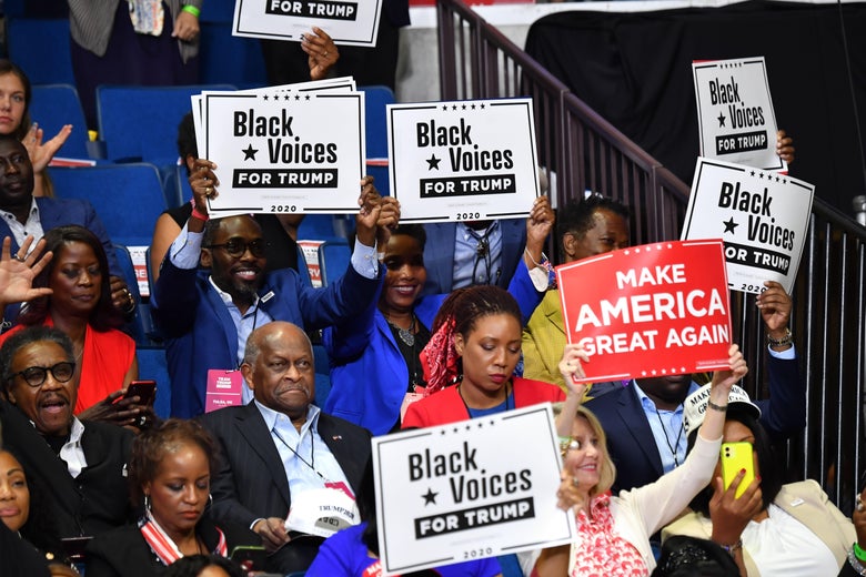 Cain sits among a group of fellow maskless Trump supporters who hold up "Black Voices for Trump" signs.