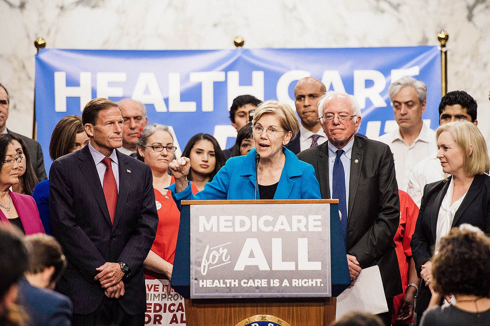 Elizabeth Warren at a podium that says, "Medicare for all," with Bernie Sanders and other senators around her.