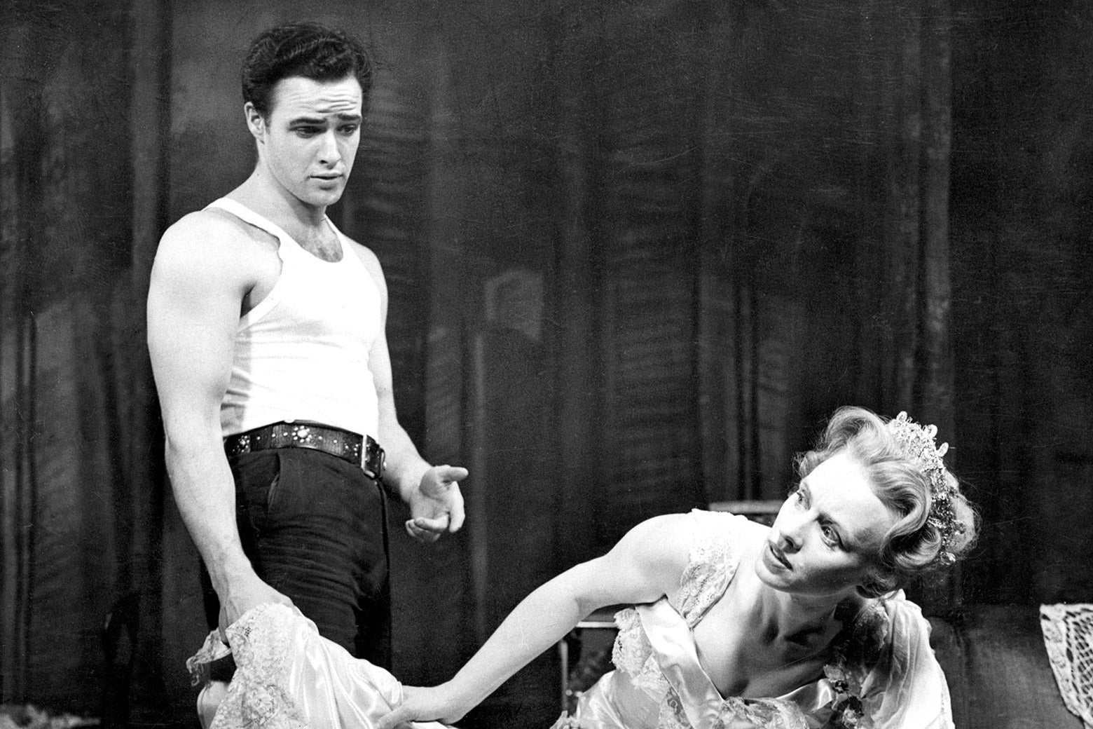 Brando as Stanley Kowalski in a white tank top and jeans stands near Tandy as Blanche DuBois in a tiara and a lacy dress sitting in a chair and leaning over to look to the side