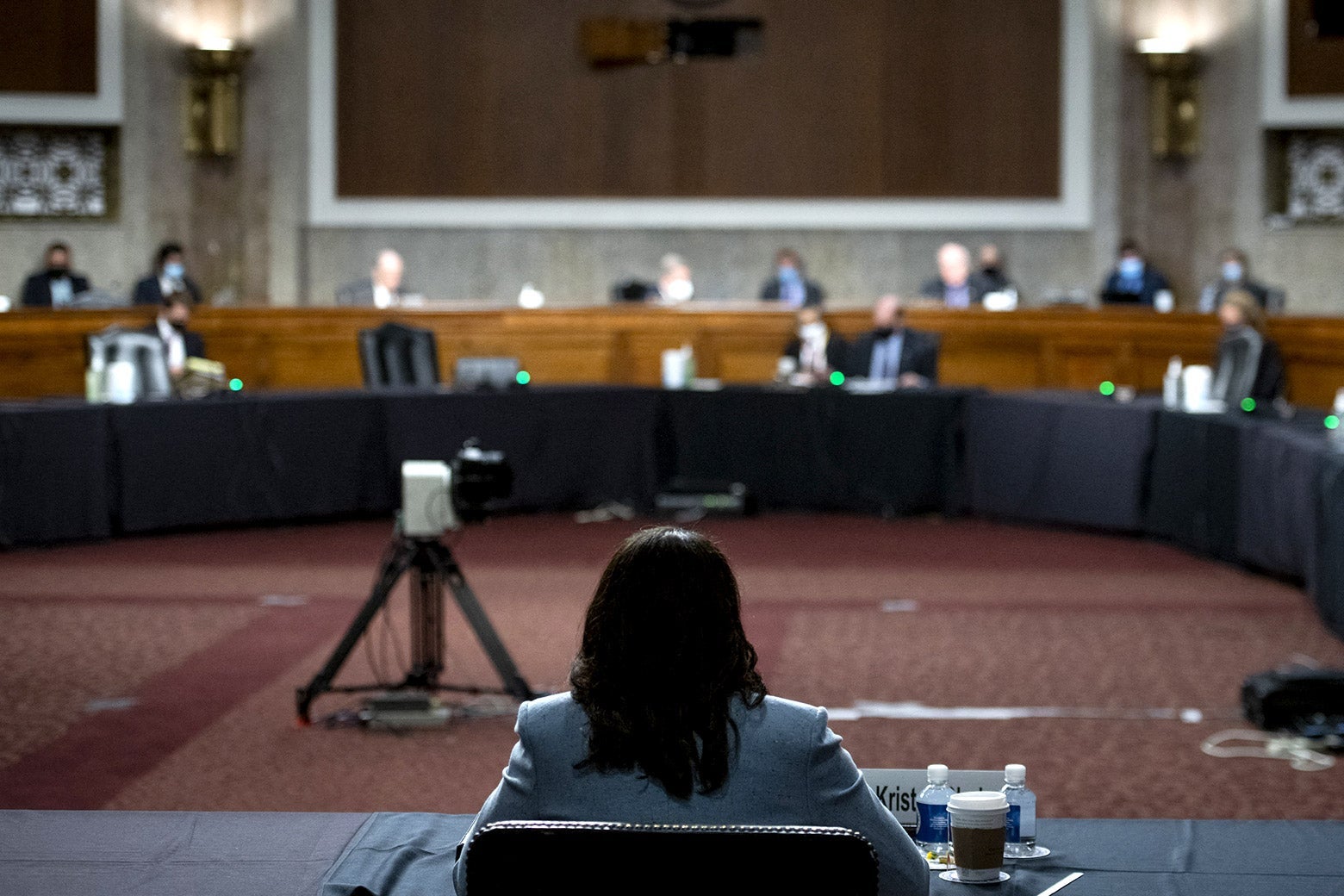 Kristen Clarke's back is seen as she sits a table inside a hearing room. Ahead in the distance, senators can be seen sitting at desks.