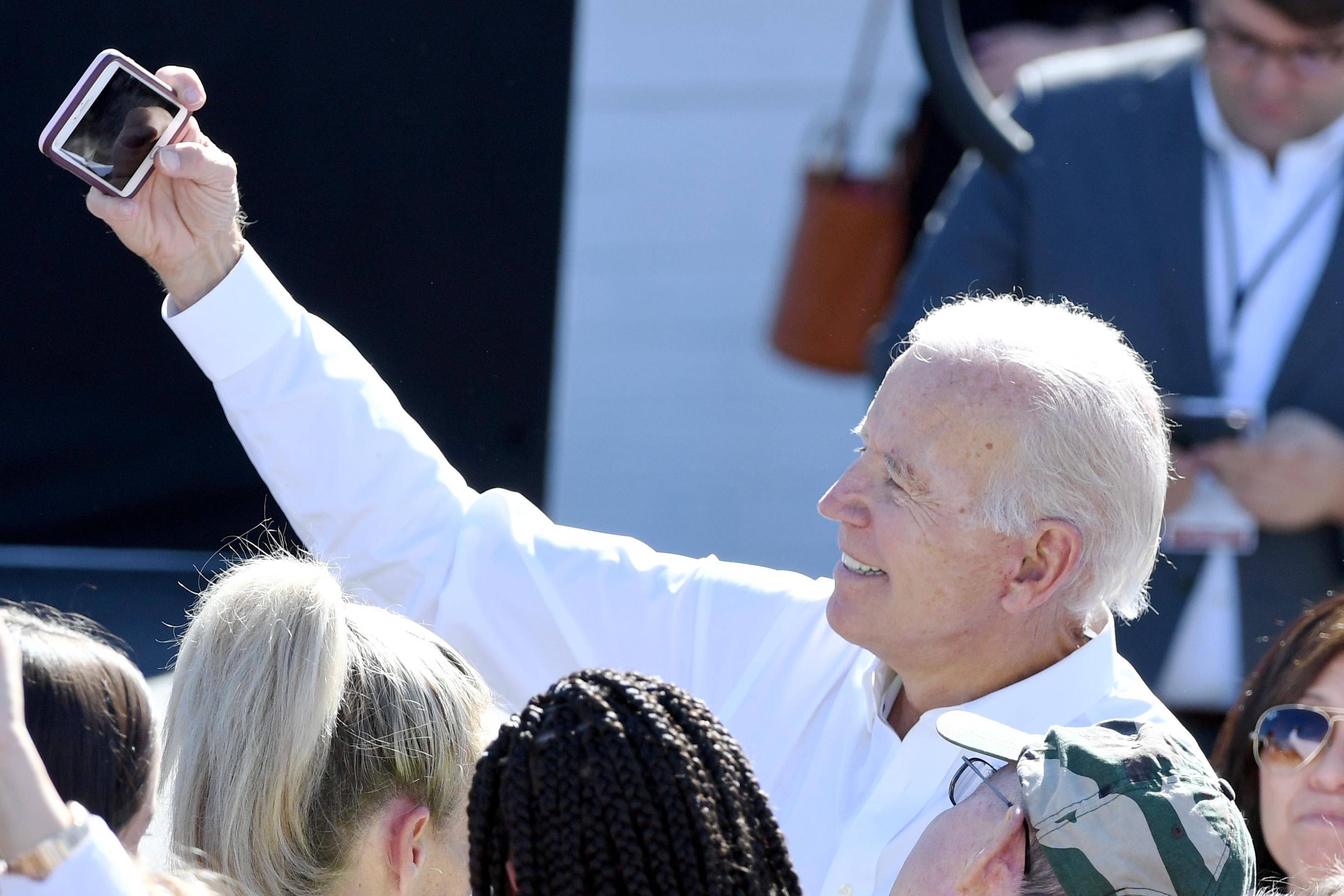 Biden raises his arm, holding a phone, to take a selfie in a crowd of people.