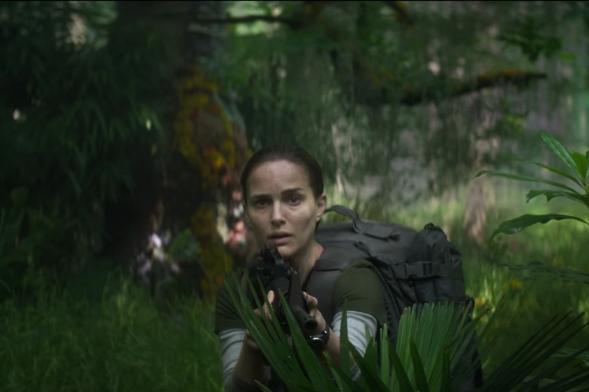Natalie Portman stands amid foliage holding a machine gun
